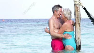 Senior Couple Embracing In The Sea Next To Hammock