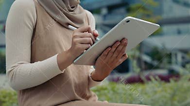 Closeup view of Malaysian woman in hijab writes with pen on tablet in city park