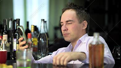 young man in a shirt is sitting in a bar drinking whiskey and reading a message on a smartphone with joy on his face.
