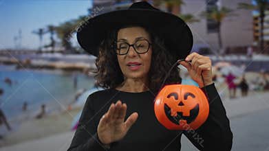 Middle age hispanic woman holds pumpkin bucket and makes a calm hand gesture at beach; serenity reassurance