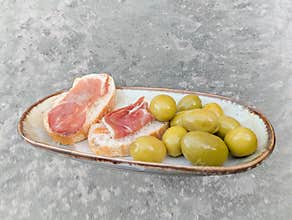 Delicious sliced bread topped with cured meat and served with green olives on a rustic plate