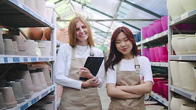 A portrait of two multiracial female employees in aprons standing between store shelves, holding clipboards, smiling