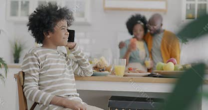 Happy African American boy making mobile call talking while parents mother and father using smartphone in kitchen at