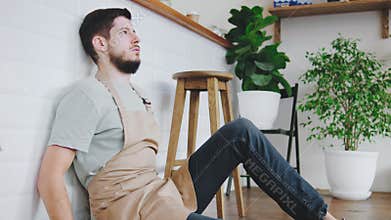 A barista guy sits on the floor of a cozy cafe, exhausted and trying to rest from his hard work. The warm and inviting