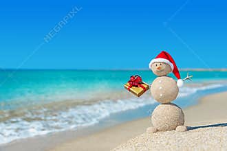 Smiley sandy snowman at beach in christmas hat with golden gift.