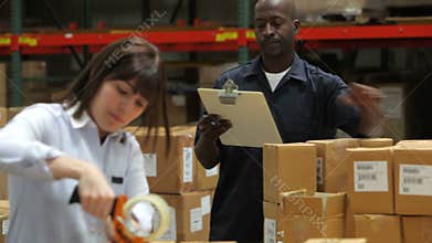 Worker Checks Clipboard As Colleague Seals Boxes
