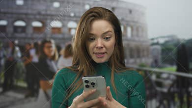 Young caucasian woman holding smartphone with both hands in front of coliseum building while looking at screen; amazement