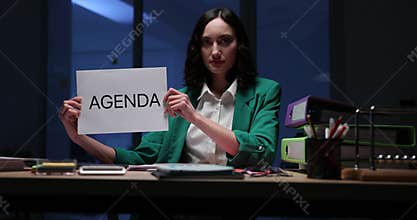Woman office worker sits at desk and holds sign with word AGENDA