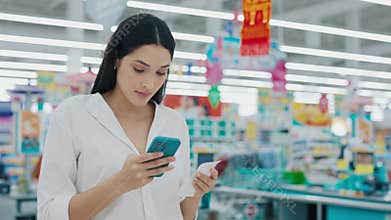 Woman Calculating All Items On Receipt By Reviewing Her List On Phone Standing Inside Supermarket