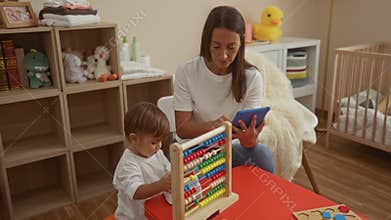 Woman sitting with son in bedroom using tablet near cradle, toys, and abacus, highlighting indoor family bonding