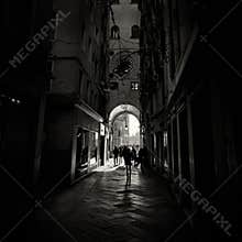 Silhouetted figures walk through a narrow, dark historic alleyway in Venice, moving towards a bright, St. Mark place illuminated