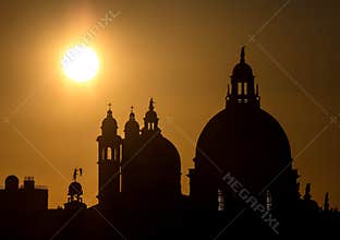Majestic domes and spires of historic Venetian architecture cast in silhouette against a dramatic, glowing golden sun at sunset