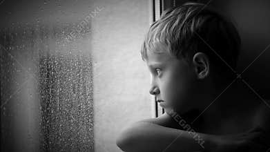 Alone little boy looks raindrops through window glass