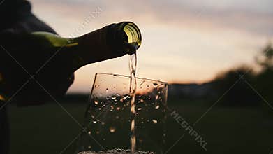 hand pours crisp white wine into a glass as sun sets, illuminating vibrant hues