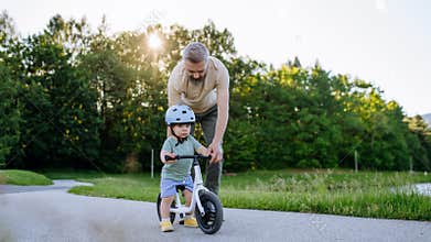 Father teaching toddler son to ride balance bike while wearing safety helmet.