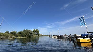 Calm canal with moored boats beside reed lined shoreline and open blue