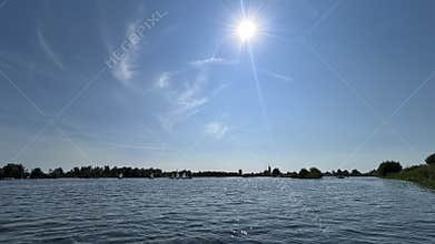 Bright sun over wide river with small sailboats, blue sky, wispy clouds
