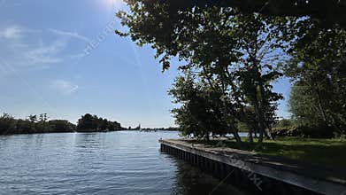 Sunny lakeside view with calm water, leafy trees, wooden dock, wide blue