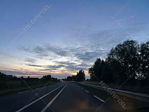 Curving highway at dusk beneath wide blue sky with glowing clouds, dark