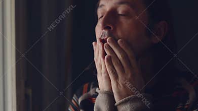 Adult caucasian sad man standing by the window of his living room and crying in despair