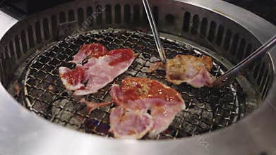 Korean barbecue grill with marinated pork slices cooking on a metal wire mesh. Metal tongs flipping meat over hot charcoal in a