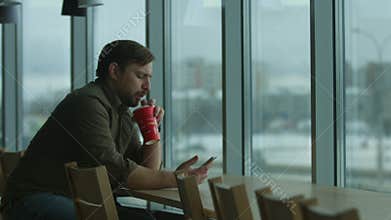 Serious man writes a message and drinks, by a large window.