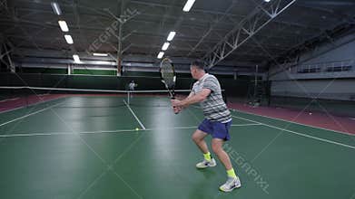 Back view of man dressed in white sport outfit serving and returning bouncing balls with racket. Professional sport