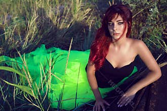 Beautiful red haired woman in black corset and long tail green veiling skirt leaning on the shabby upside down wooden boat