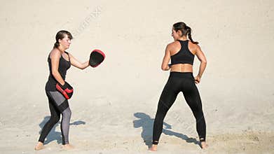 Two athletic, young women in black fitness suits are engaged in a pair, work out kicks, on a deserted beach, against a