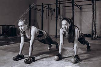 Two fit women in gym doing fitness exercises with dumbbells staying in plank pose, black and white