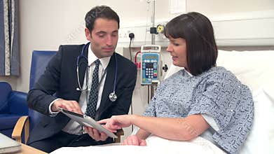 Doctor Sitting By Female Patient's Bed Discussing Notes