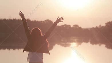 Young girl slowly raises her hands up enjoying sunrise on the river, then turns around and looks at the camera