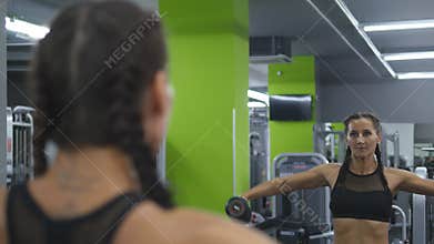 Young pretty woman trains in the gym lifting and lowering dumbbells in front of the mirror. Sport fitness girl