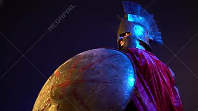 Roman gladiator in leather armor, helmet and red raincoat is covered with a round shield and strikes with a sword, slow