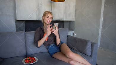 Blond woman holds mobile phone and eats ripe strawberry, sitting on soft sofa in living room with gray walls in
