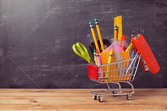 Shopping cart with school supplies over chalkboard background. Back to school sale concept