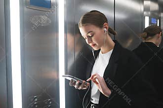 Smiling businesswoman is using her smartphone with headphones in the elevator. Cheerful office worker is texting on the phone and