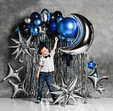 Kid boy stands on background of silver and blue metallic balloons for birthday party holding hand up and smiles