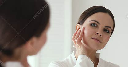 Beautiful young 20s woman applying collagen moisturizer on cheek.