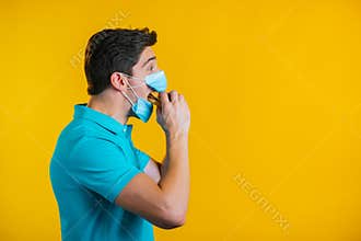 Young man in protective medical mask eating burger on yellow studio background. Guy makes fun, shows how to live during