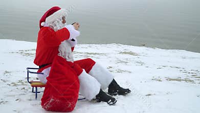 Santa Claus sits with a bag of presents on the snowy shore of a lake and drinks mulled wine. Drunk Santa Claus