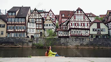 Long-hair woman in yellow coat with backpack sitting sideways near water against a background of traditional half
