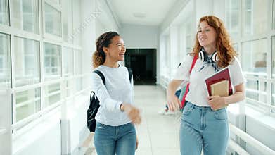 Girls friends walking in school hall holding books chatting doing high-five