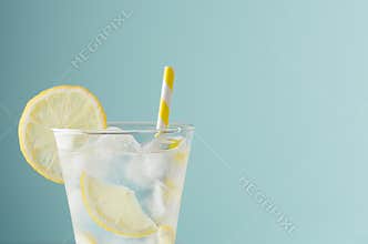 Cold shine sparkling refreshing drink for party with lemon slices, ice cubes, tonic, yellow straw in elegant glass on mint color.