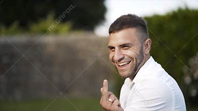 Close-up view of handsome bridegroom with brown hair and beard in white shirt looking at the camera and smiling in park