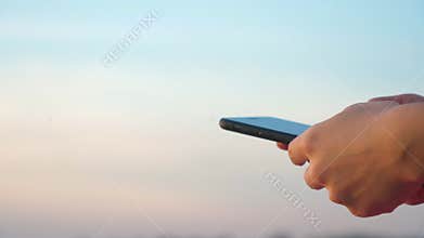 Woman`s hand using mobile phone with a clear sky in the background.