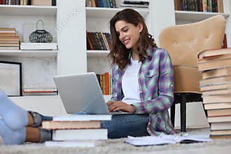 Smiling young female sitting on the floor in the living room student studying at home working with her laptop.