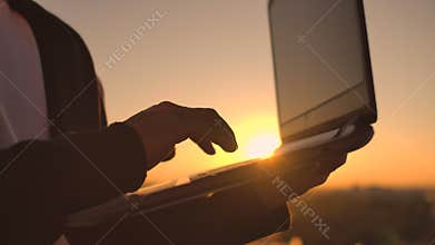 Close-up: a programmer's hand typing on a laptop keyboard at sunset overlooking the roof. A businessman works remotely