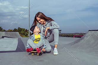 Woman mother encourages and motivates child on skateboard, little boy 3-5 years old, summer sports field in city, autumn