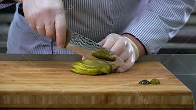 Chef in rubber gloves cuts pickled cucumber with knife on wooden bamboo cutting Board.